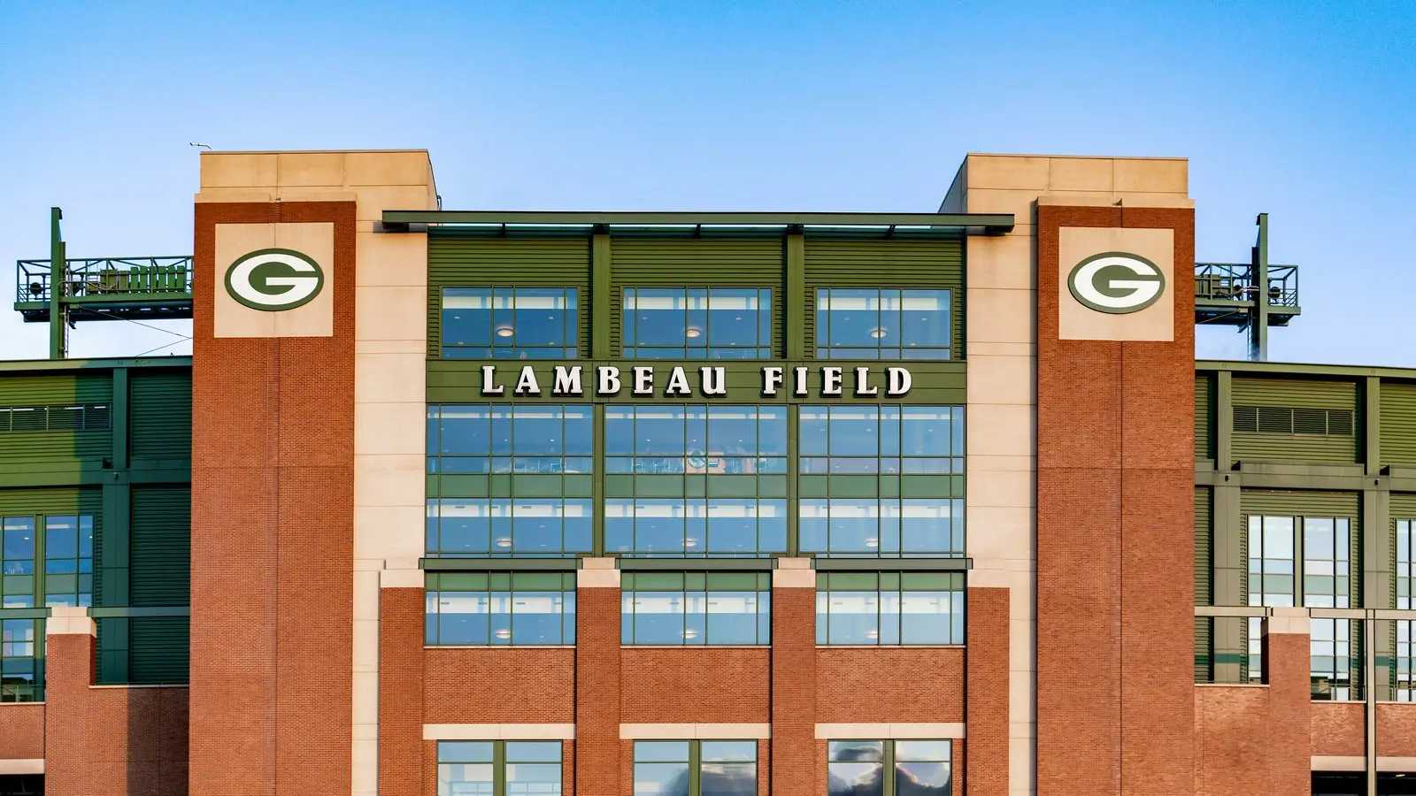 Brick and glass facade of Lambeau Field in Green Bay, Wisconsin, with the stadium name and two Green Bay Packers G logos under a blue sky