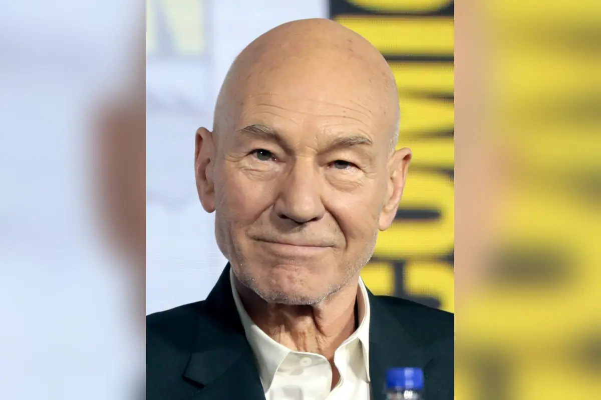 Patrick Stewart, a bald older man in a dark jacket and white shirt, smiling on a panel stage