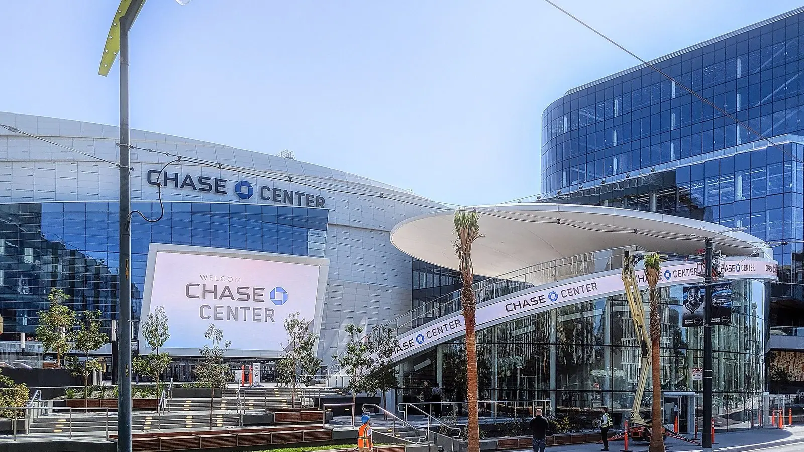 Exterior of Chase Center in San Francisco with large Chase Center signs, a curved white canopy and glass office towers under a clear blue sky